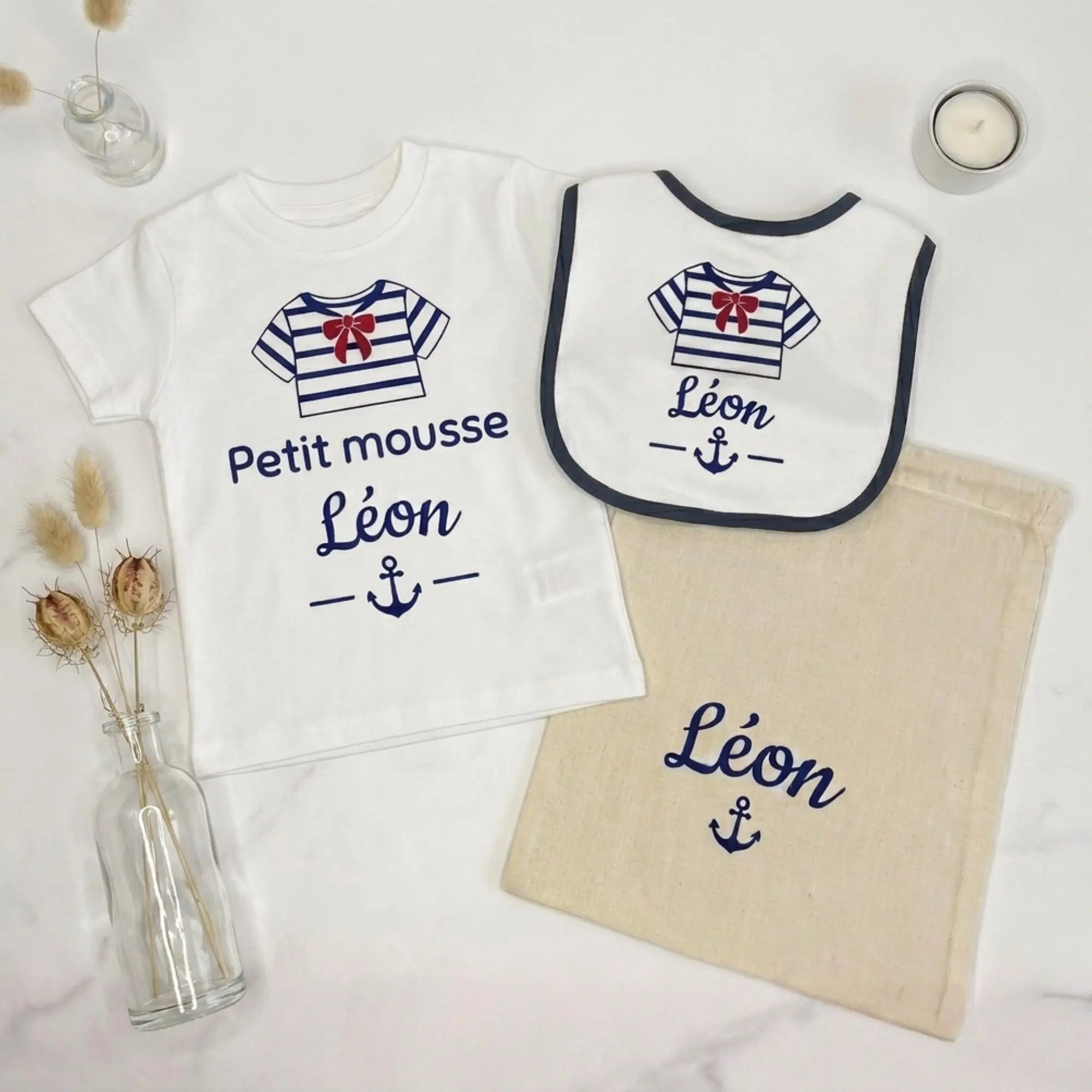 Coffret personnalisé enfant Petit Mousse composé d'un t-shirt blanc, d'un bavoir et d'un petit sac en toile. Graphisme marinière bleu marine, prénom Léon et petit nœud rouge, présenté sur un fond blanc épuré.