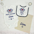Coffret cadeau bébé personnalisé Petit Mousse comprenant un body blanc, un bavoir et un pochon en tissu. Flocage bleu marine au prénom de Léon avec motif de marinière et nœud rouge, posé sur un fond épuré en marbre blanc.