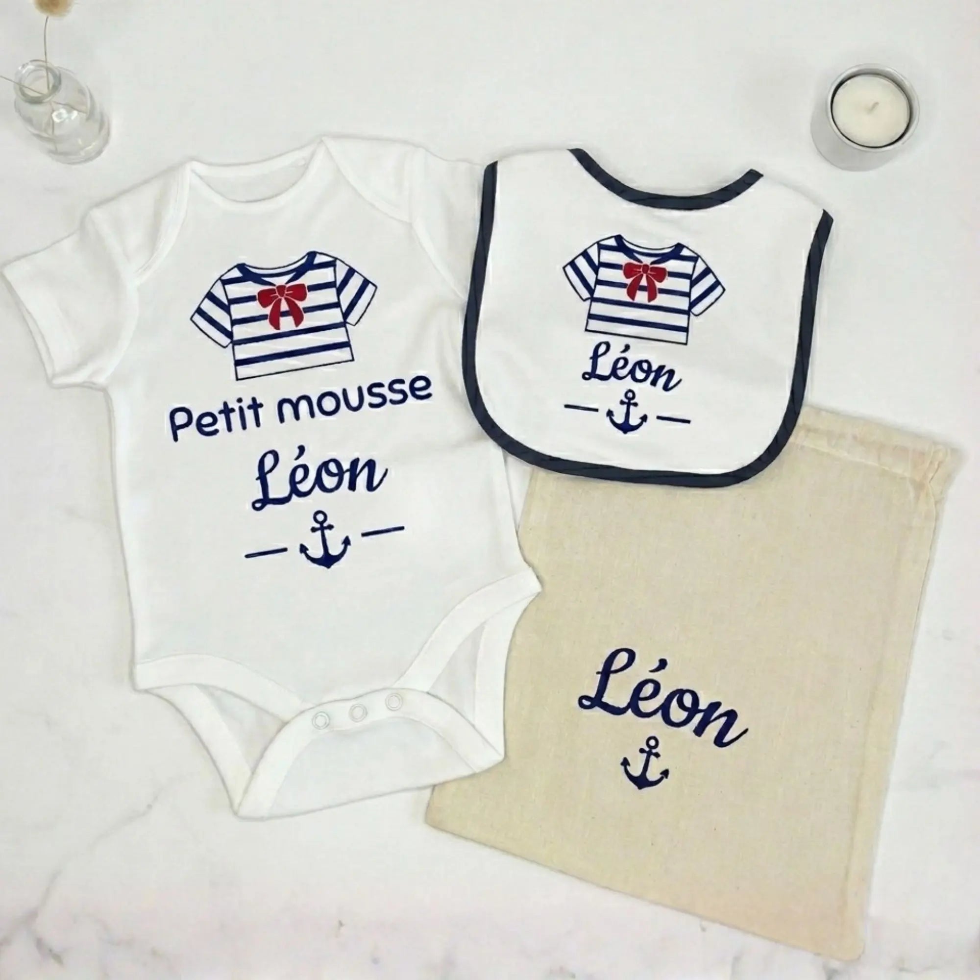 Coffret cadeau bébé personnalisé Petit Mousse comprenant un body blanc, un bavoir et un pochon en tissu. Flocage bleu marine au prénom de Léon avec motif de marinière et nœud rouge, posé sur un fond épuré en marbre blanc.