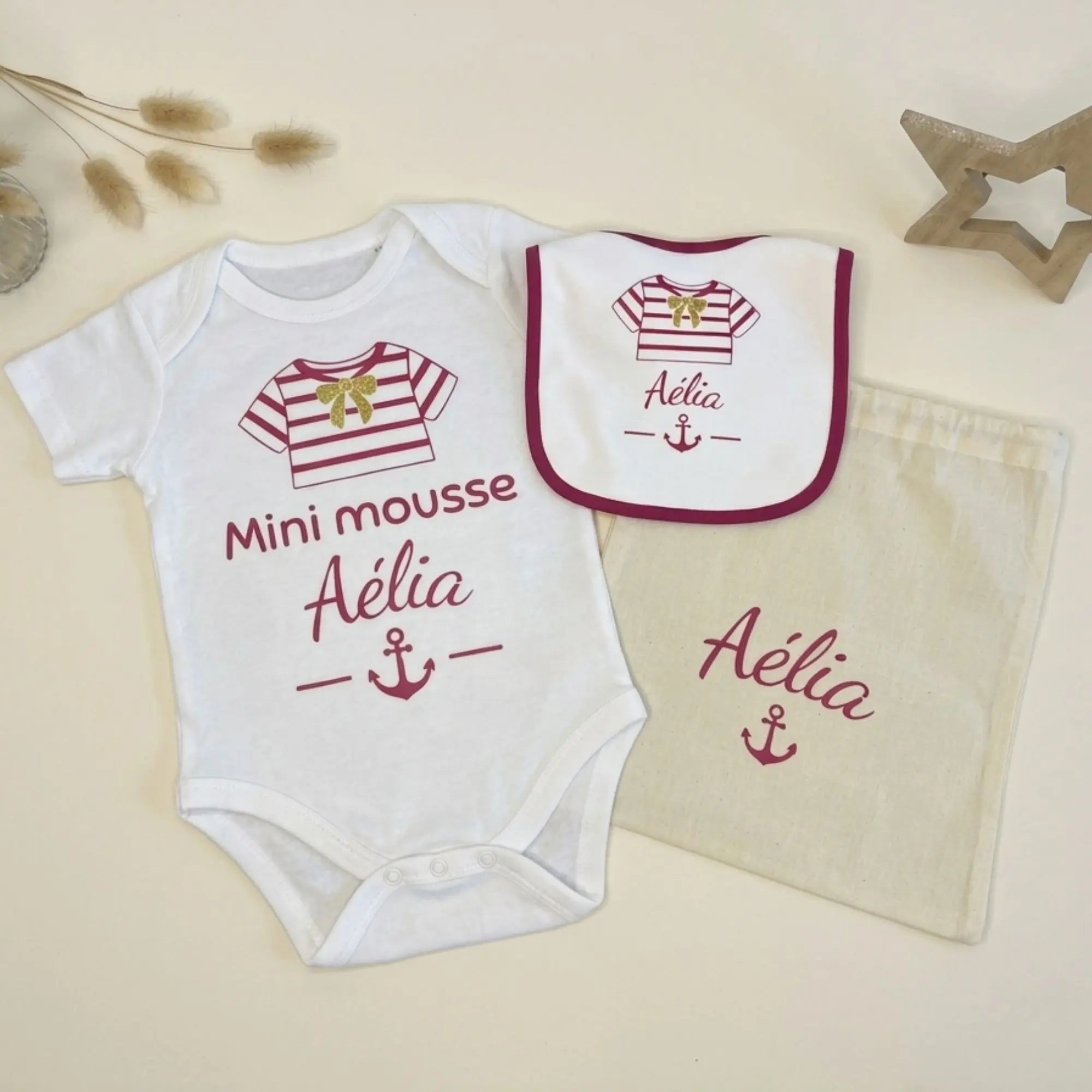 Coffret naissance personnalisé Mini Mousse de la marque Bébé d'Armor, comprenant un body blanc, un bavoir assorti et un pochon en coton beige, tous floqués au prénom d'Aélia avec un motif de marinière rose et une ancre marine, posés sur un fond épuré.