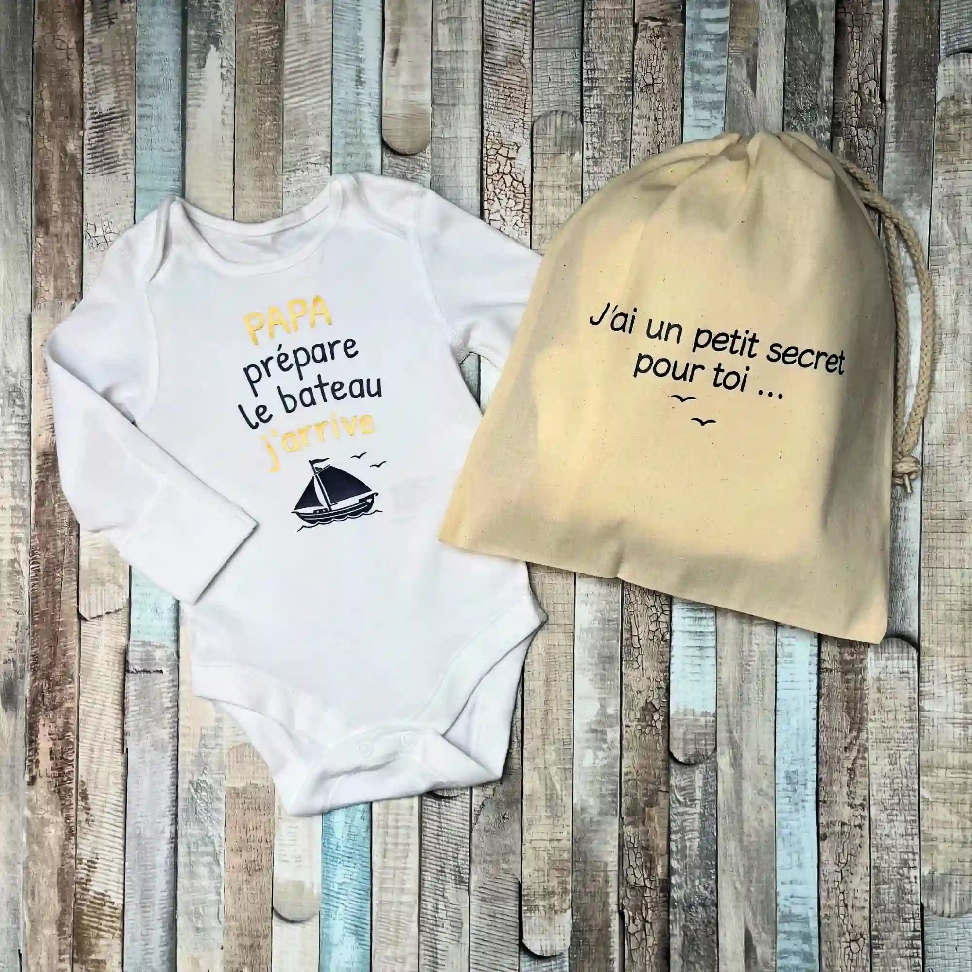 Body bébé blanc manches longues personnalisé "Papa prépare le bateau j'arrive" à côté d'un pochon en tissu "J'ai un petit secret pour toi"