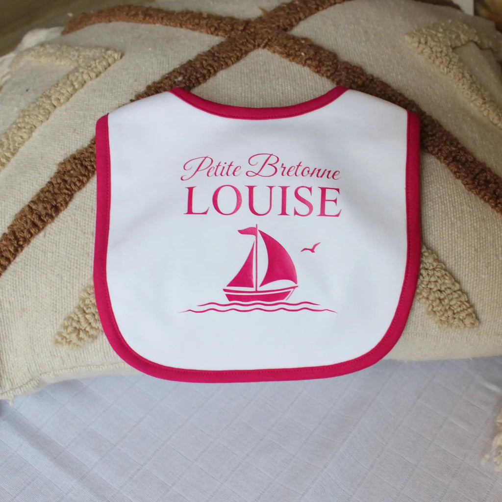 Bavoir blanc pour bébé fille avec un liseré rose fuchsia, personnalisé avec l'inscription Petite Bretonne Louise et un dessin de voilier rose. Posé sur un coussin décoratif.