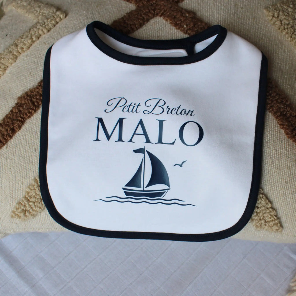 Bavoir blanc pour bébé garçon avec un liseré noir, personnalisé avec l'inscription Petit Breton Malo et un dessin de voilier marine. Posé sur un coussin décoratif.