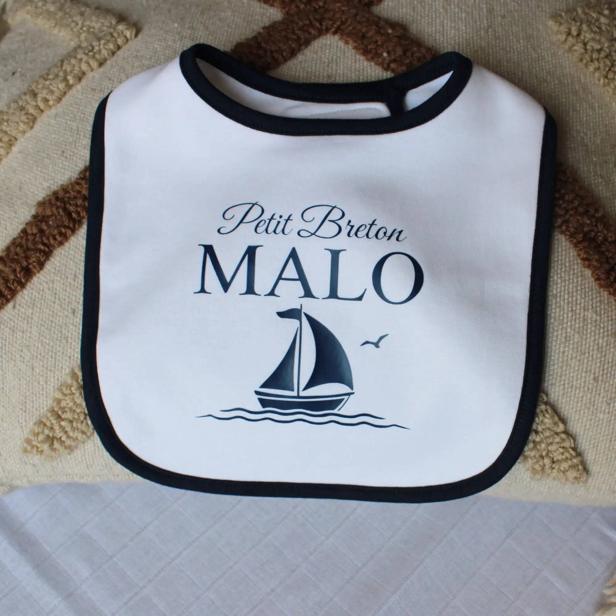 Bavoir blanc pour bébé garçon avec un liseré noir, personnalisé avec l'inscription Petit Breton Malo et un dessin de voilier marine. Posé sur un coussin décoratif.