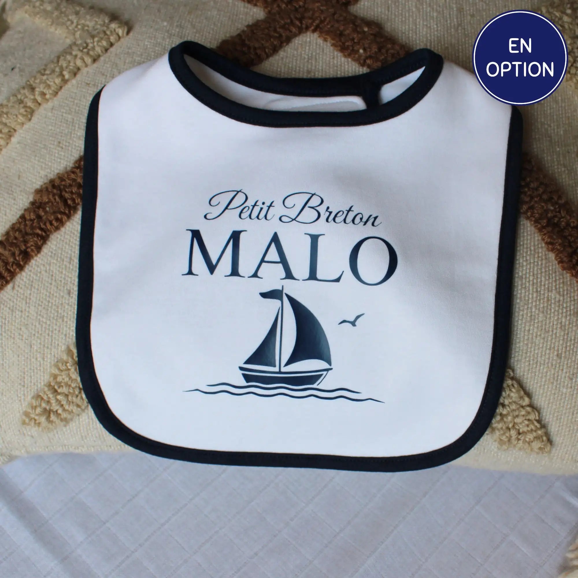 Bavoir blanc pour bébé garçon avec un liseré noir, personnalisé avec l'inscription Petit Breton Malo et un dessin de voilier marine. Posé sur un coussin décoratif.