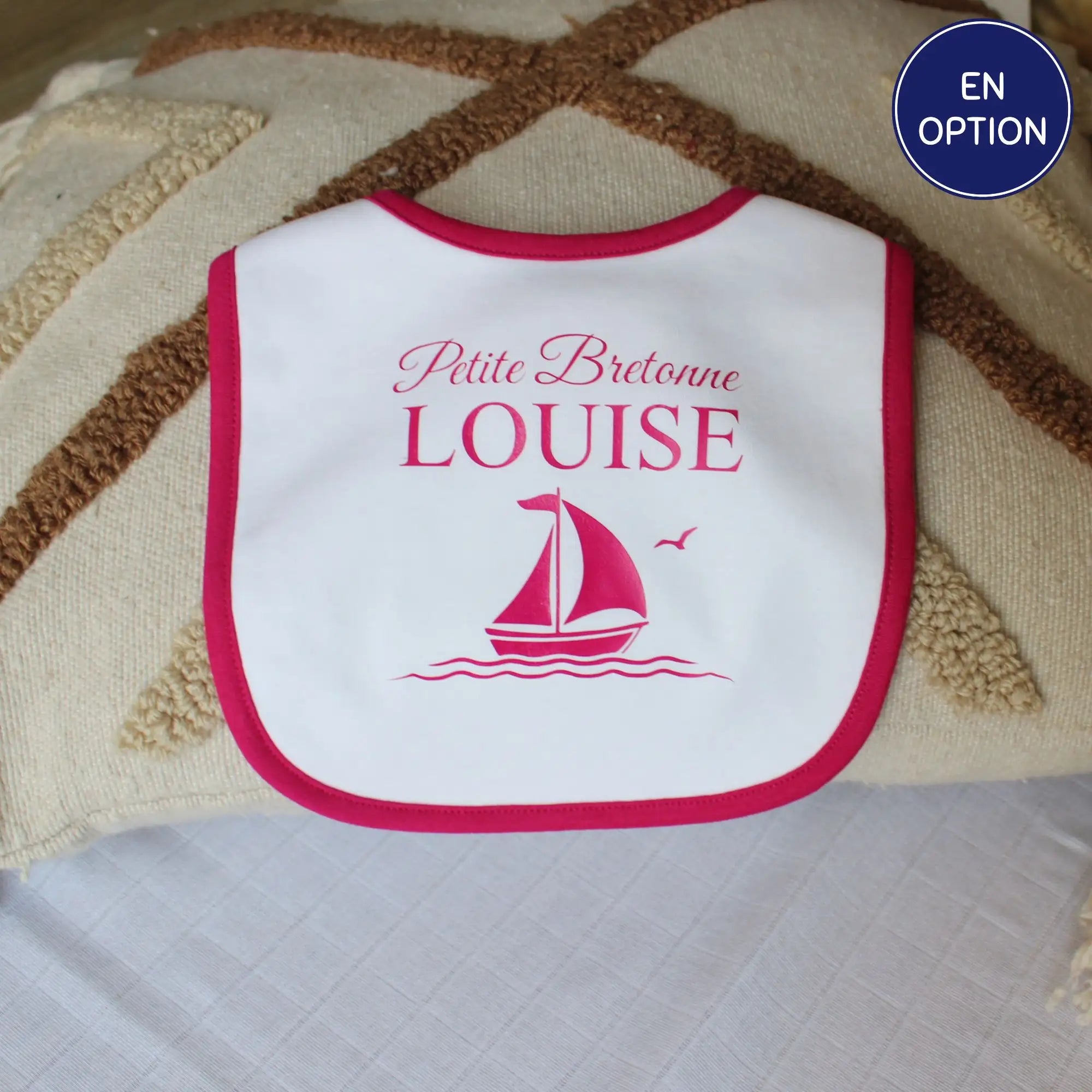 Bavoir blanc pour bébé fille avec un liseré rose fuchsia, personnalisé avec l'inscription Petite Bretonne Louise et un dessin de voilier rose. Posé sur un coussin décoratif.