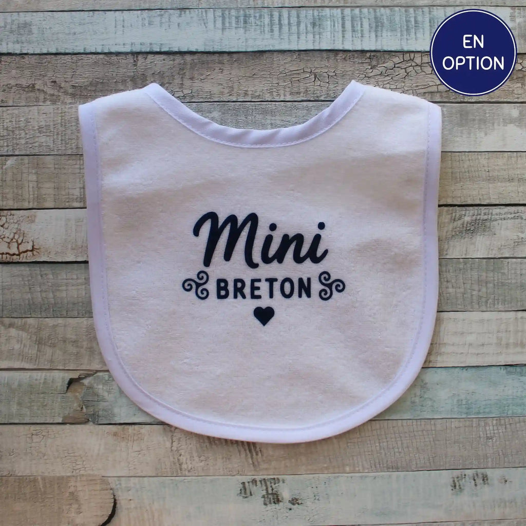 Un bavoir blanc pour bébé orné de l'inscription "Mini Breton" en écriture cursive bleue, entouré de motifs bretons traditionnels et d'un petit cœur bleu, le tout posé sur un fond en bois clair rustique.