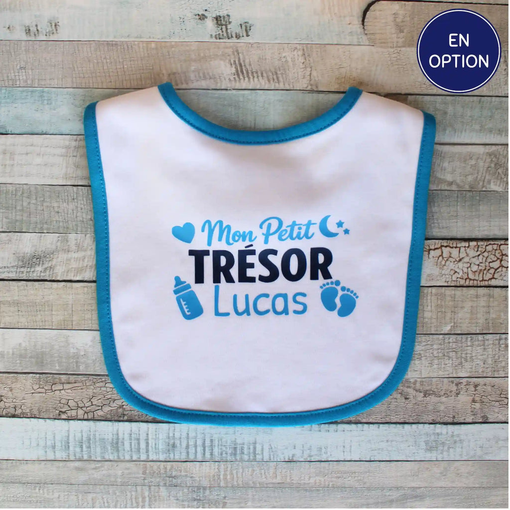 Photo à plat d'un bavoir blanc bordé de bleu vif, posé sur un parquet en bois délavé, affichant la personnalisation "Mon Petit Trésor Lucas"