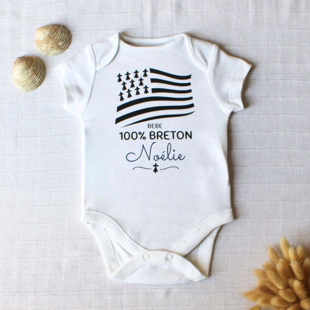 Body blanc pour bébé en coton personnalisé avec le prénom Noélie et le drapeau breton Gwenn ha du, posé à plat avec des coquillages.