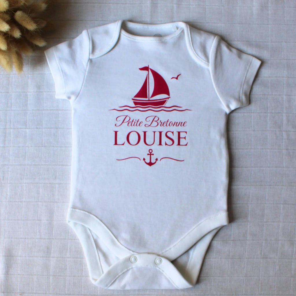 Body blanc à manches courtes pour bébé fille, personnalisé avec le prénom Louise, l'inscription Petite Bretonne, un bateau à voile et une ancre, le tout en rose framboise. Posé à plat avec une décoration florale.