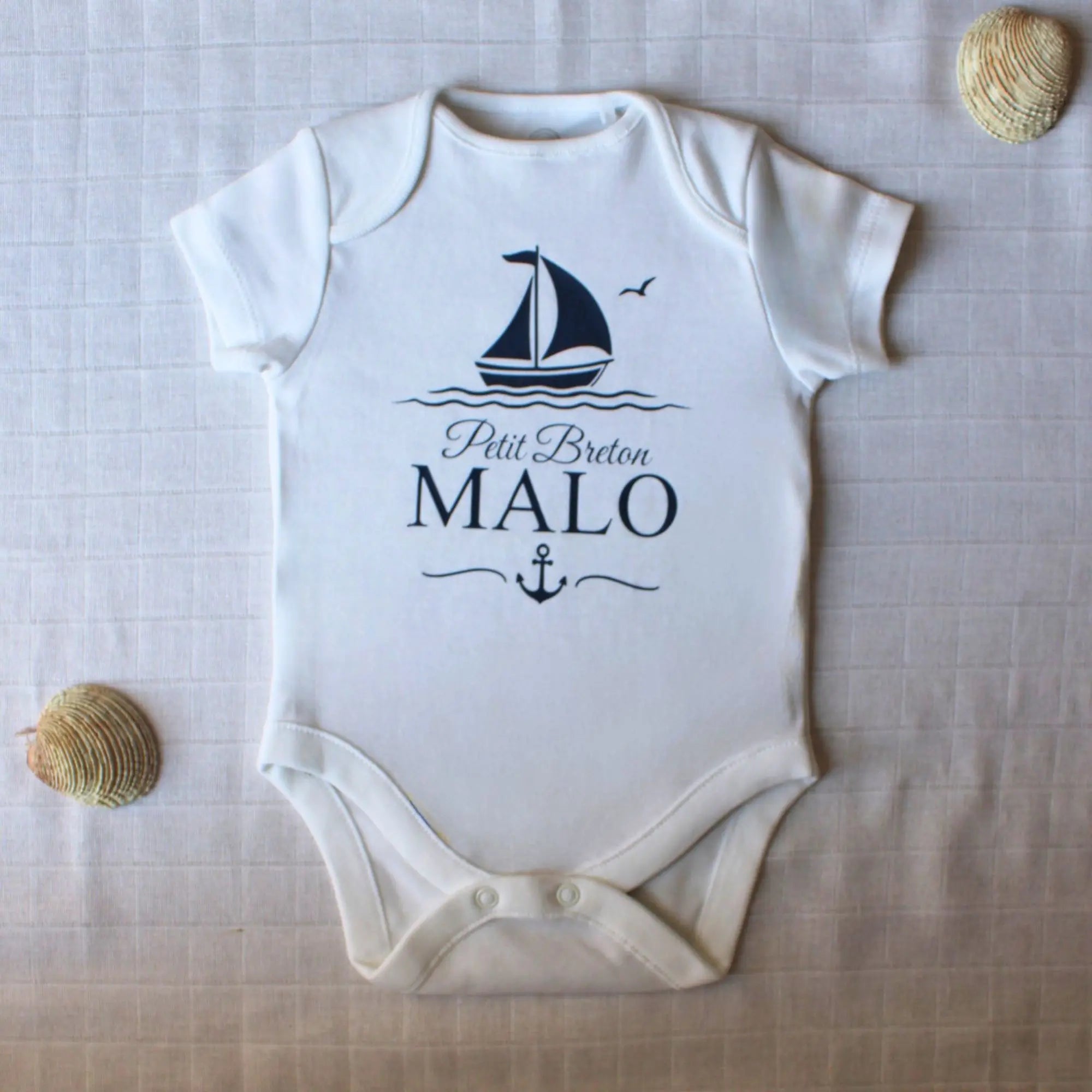 Body blanc à manches courtes pour bébé garçon, personnalisé avec le prénom Malo, l'inscription Petit Breton, un bateau à voile et une ancre, le tout en bleu marine. Posé à plat avec des coquillages.