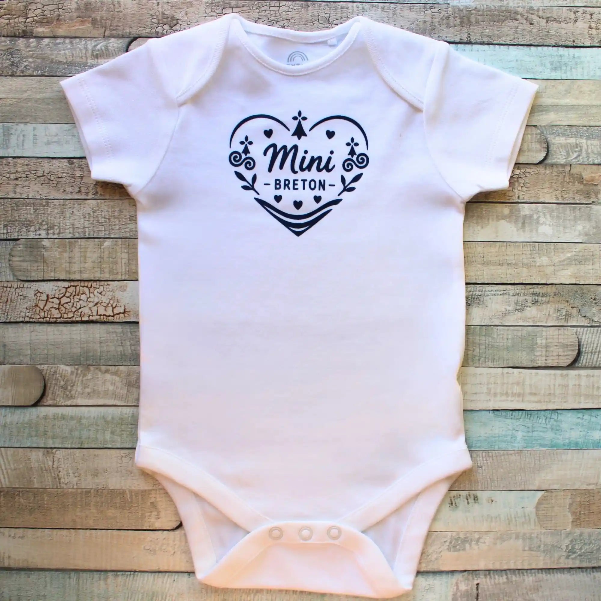 Un body blanc à manches courtes pour bébé, présentant un grand cœur central composé de motifs celtiques bleus et de l'inscription "Mini Breton" en son centre. Un badge bleu circulaire dans le coin supérieur droit indique "Disponible en manches longues". Le vêtement est posé à plat sur un fond en bois gris délavé.