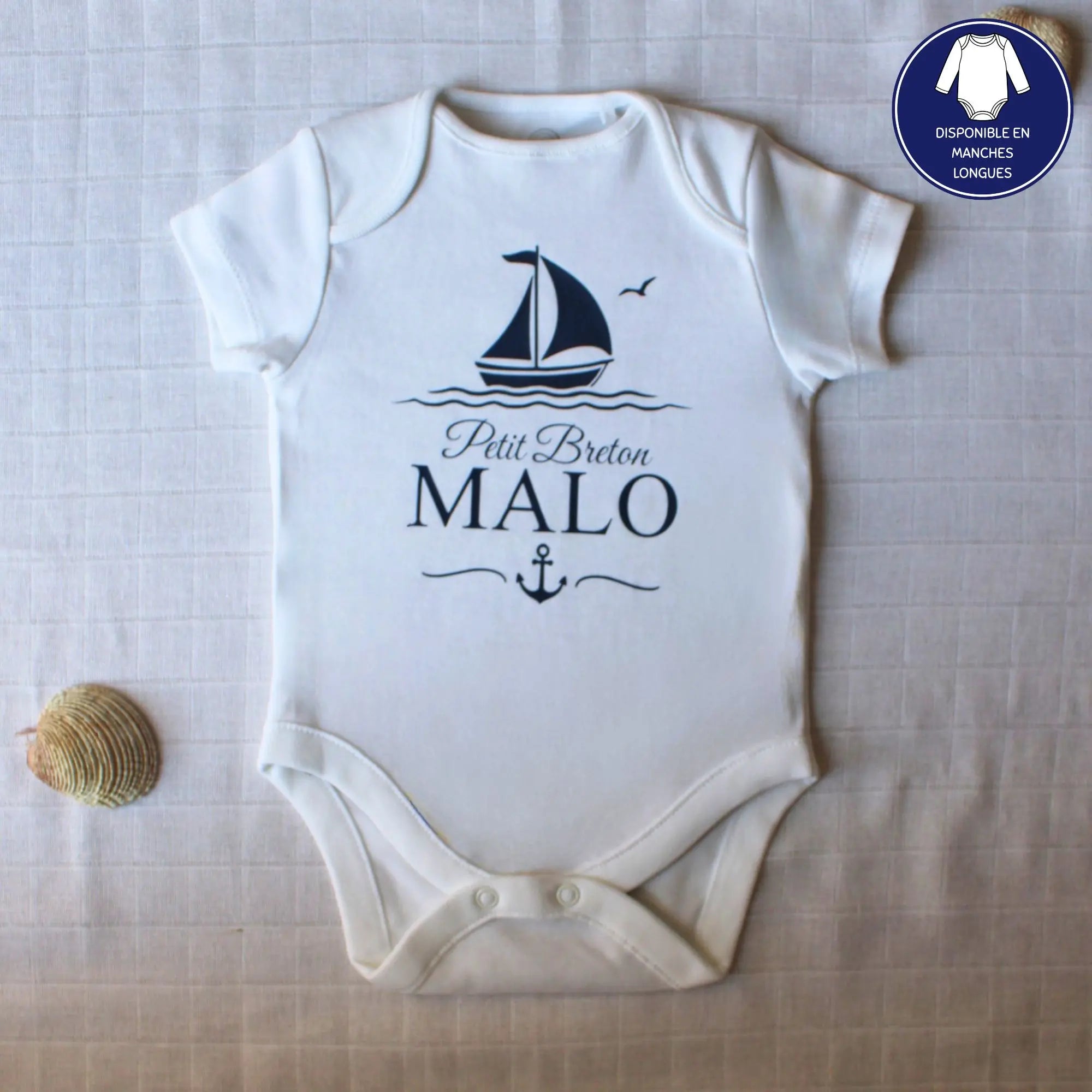 Body blanc à manches courtes pour bébé garçon, personnalisé avec le prénom Malo, l'inscription Petit Breton, un bateau à voile et une ancre, le tout en bleu marine. Posé à plat avec des coquillages.