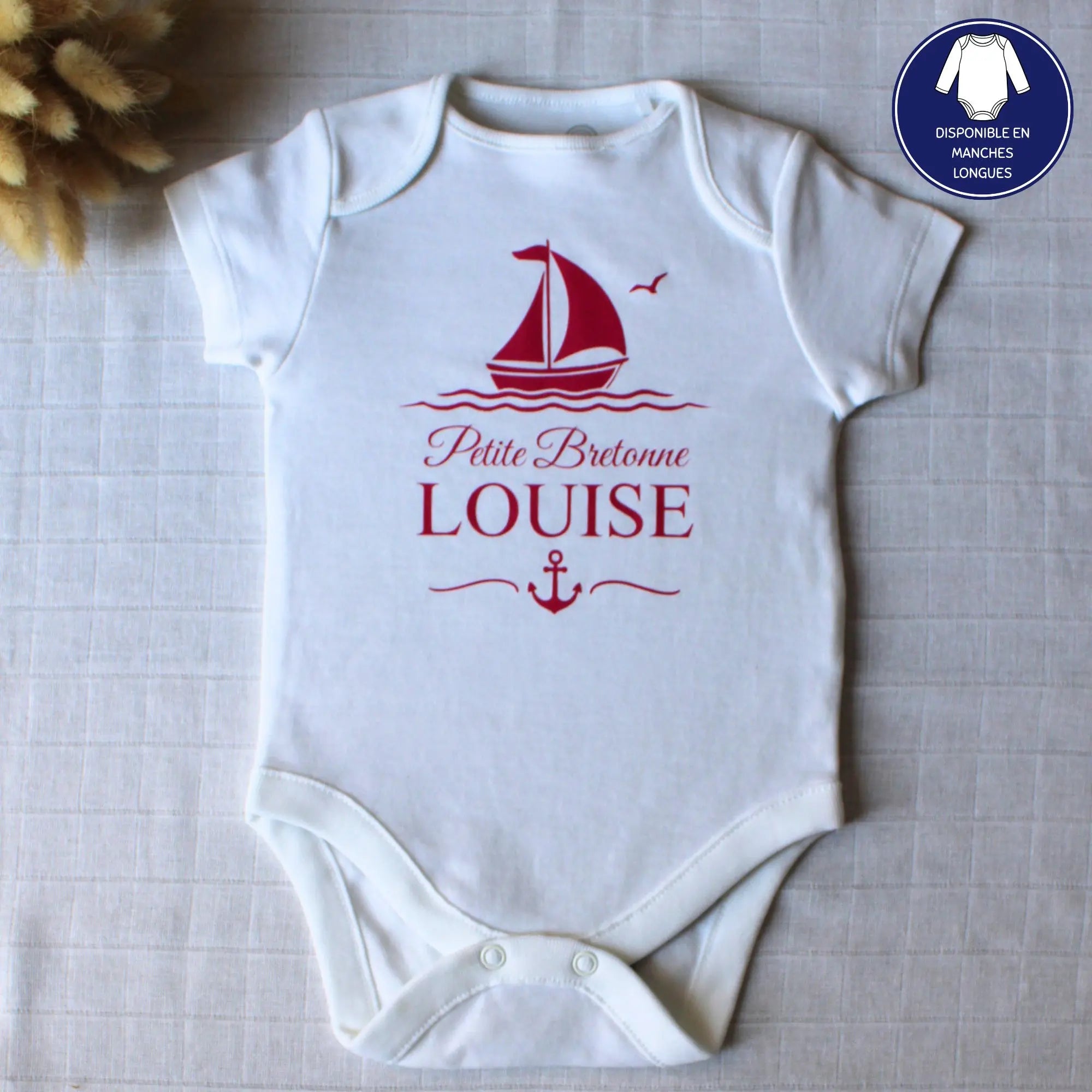 Body blanc à manches courtes pour bébé fille, personnalisé avec le prénom Louise, l'inscription Petite Bretonne, un bateau à voile et une ancre, le tout en rose fuchsia. Posé à plat avec une décoration florale.