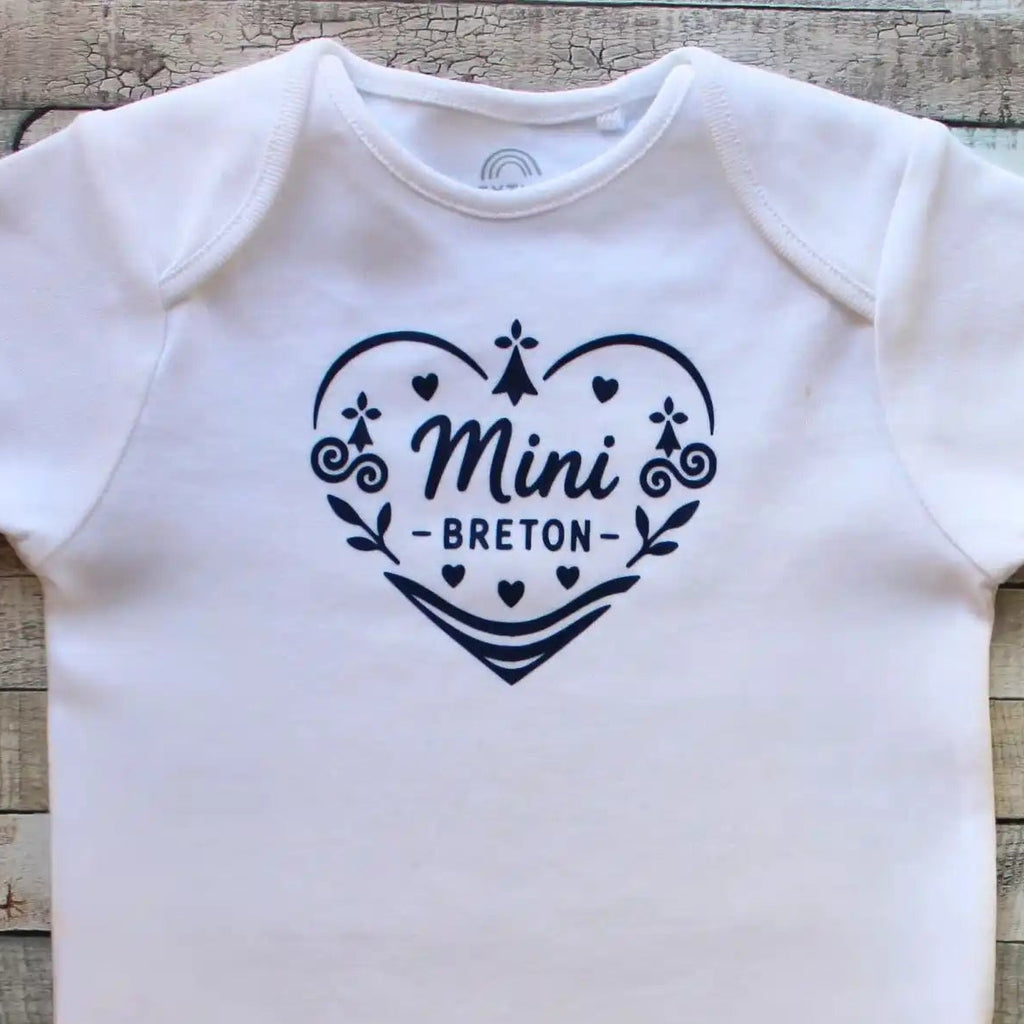Un body blanc à manches courtes pour bébé, présentant un grand cœur central composé de motifs celtiques bleus et de l'inscription "Mini Breton" en son centre. Un badge bleu circulaire dans le coin supérieur droit indique "Disponible en manches longues". Le vêtement est posé à plat sur un fond en bois gris délavé.