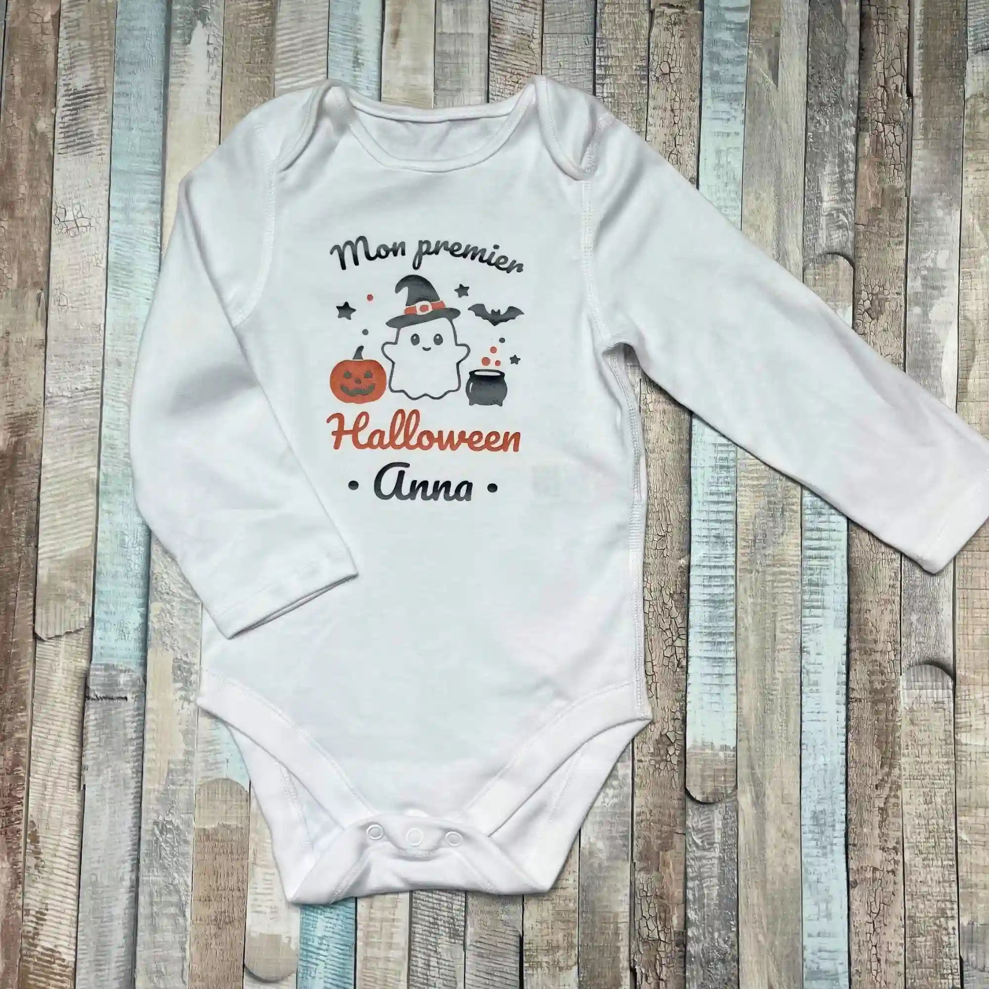 Body bébé personnalisé blanc "Mon premier Halloween Anna" avec motifs fantôme, citrouille et chaudron sur fond bois.
