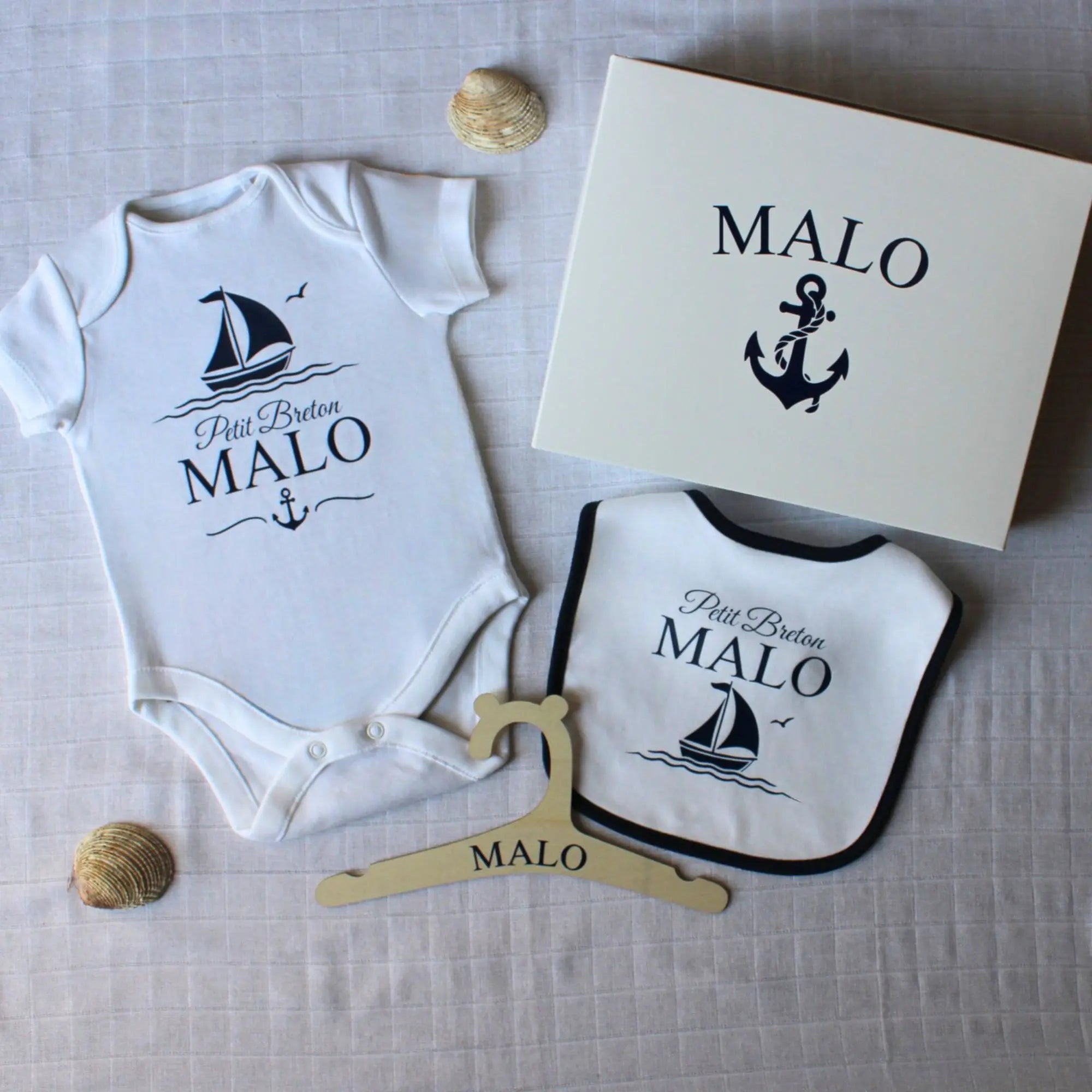 Un coffret cadeau de naissance personnalisé pour garçon prénom Malo. Le kit comprend un body, un bavoir à liseré bleu marine, un cintre en bois et une boîte cadeau, tous décorés d'un motif de bateau marine.