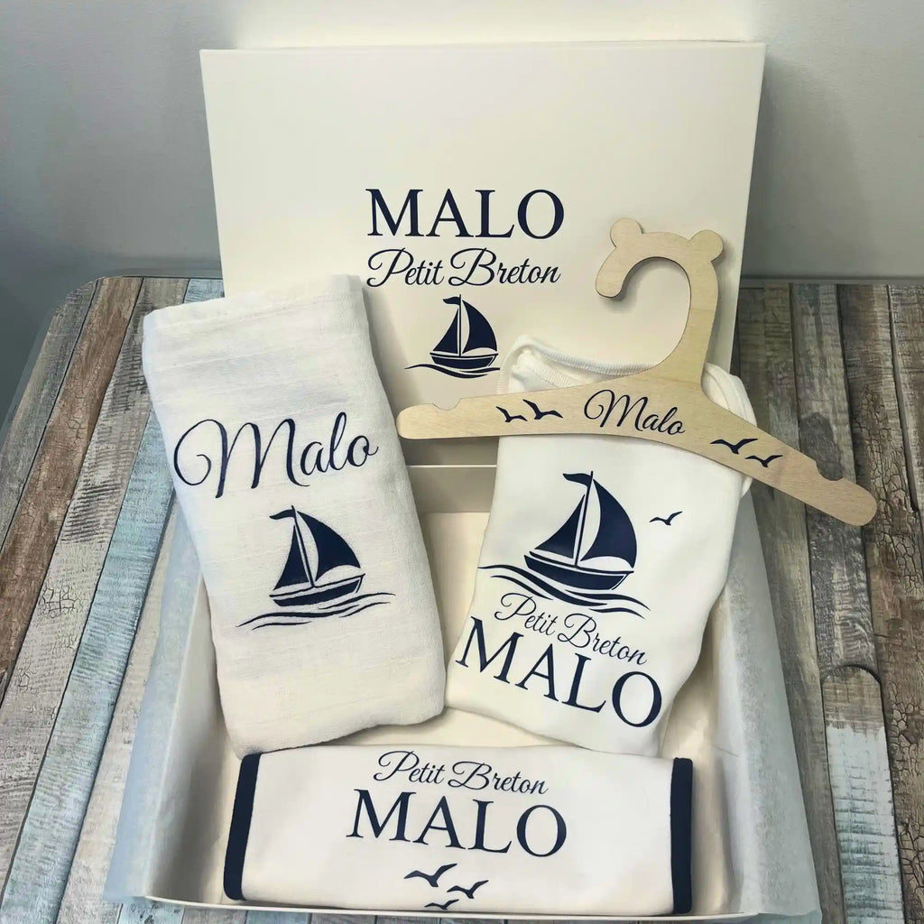 Un coffret cadeau de naissance petit breton personnalisé pour garçon prénom Malo. Le kit comprend un body, un bavoir à liseré bleu marine, un cintre en bois, un lange et une boîte cadeau, tous décorés d'un motif de bateau marine.