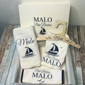 Un coffret cadeau de naissance petit breton personnalisé pour garçon prénom Malo. Le kit comprend un body, un bavoir à liseré bleu marine, un cintre en bois, un lange et une boîte cadeau, tous décorés d'un motif de bateau marine.