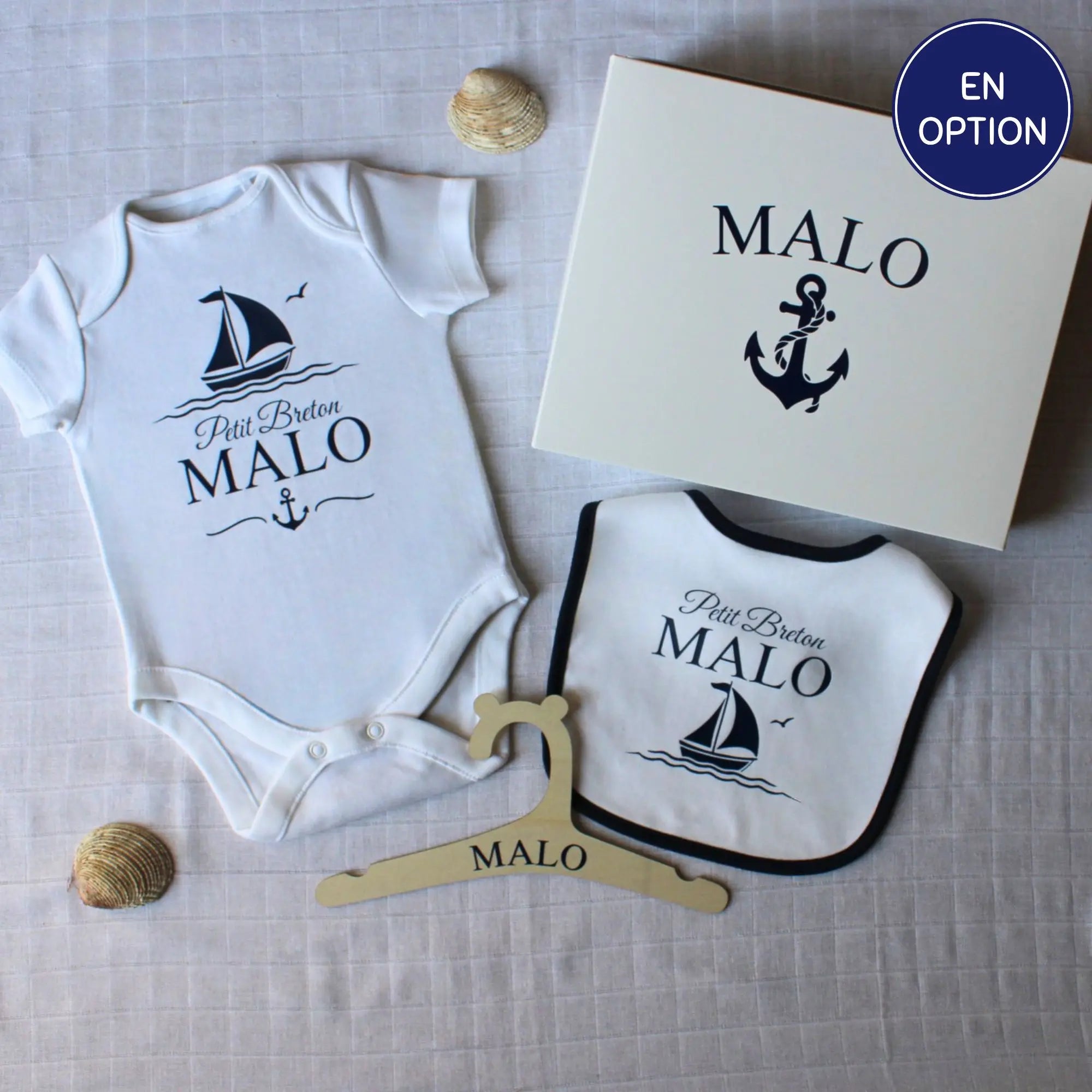 Un coffret cadeau de naissance personnalisé pour garçon prénom Malo. Le kit comprend un body, un bavoir à liseré bleu marine, un cintre en bois et une boîte cadeau, tous décorés d'un motif de bateau et ancre marine 