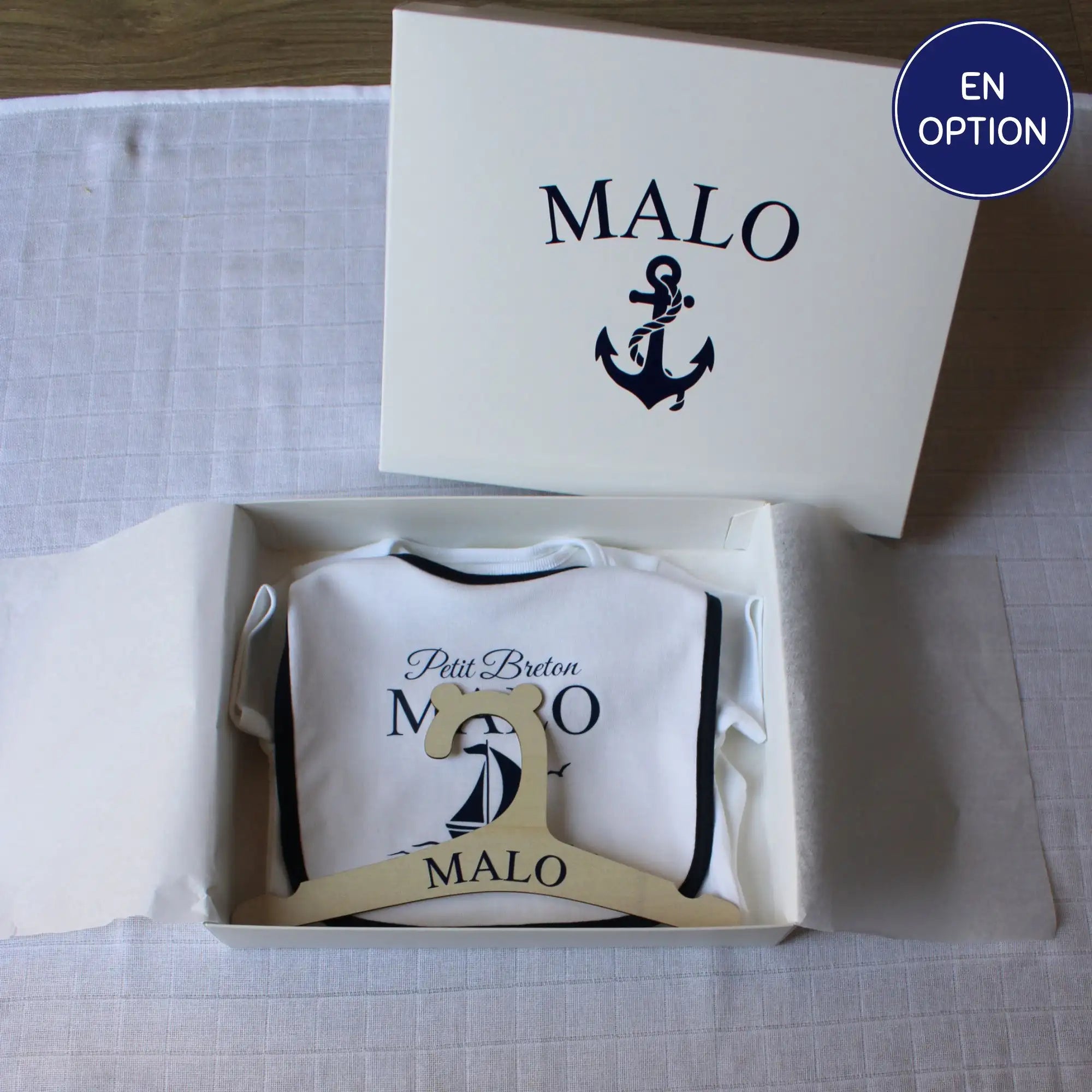Un coffret cadeau de naissance personnalisé pour garçon prénom Malo. Le kit comprend un body, un bavoir à liseré bleu marine, un cintre en bois et une boîte cadeau, tous décorés d'un motif de bateau marine. et ancres marines