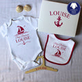 Un coffret cadeau de naissance personnalisé pour fille prénom Louise. Le kit comprend un body, un bavoir à liseré rose framboise, un cintre en bois et une boîte cadeau, tous décorés d'un motif de bateau et d'ancre