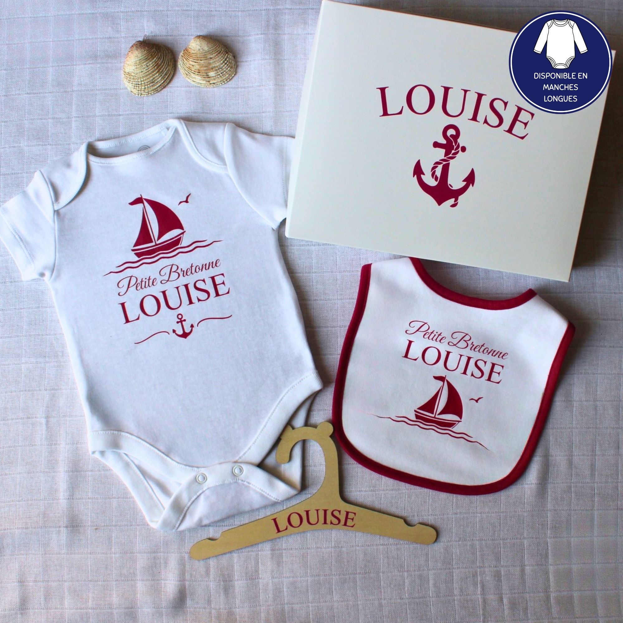 Un coffret cadeau de naissance personnalisé pour fille prénom Louise. Le kit comprend un body, un bavoir à liseré rose framboise, un cintre en bois et une boîte cadeau, tous décorés d'un motif de bateau et d'ancre
