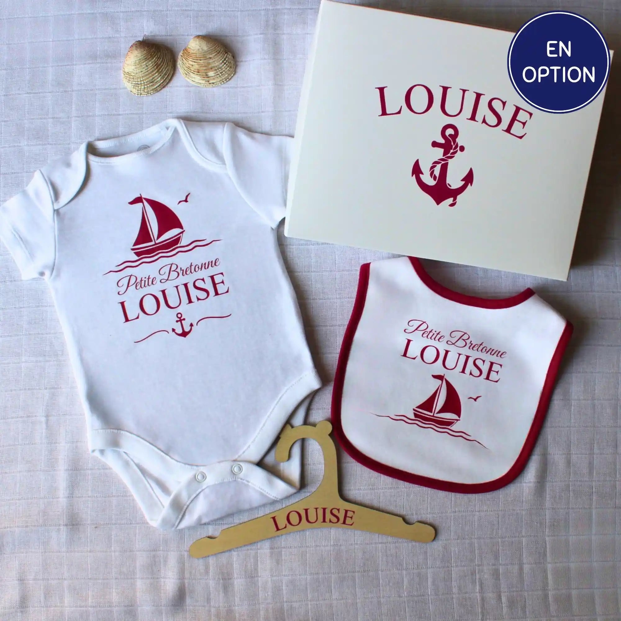 Un coffret cadeau de naissance personnalisé pour fille prénom Louise. Le kit comprend un body, un bavoir à liseré rose framboise, un cintre en bois et une boîte cadeau, tous décorés d'un motif de bateau marine. et ancres marines