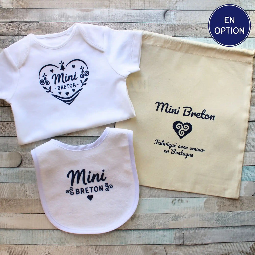 Un coffret cadeau complet présenté sur un fond en bois rustique. Il comprend un body blanc pour bébé avec le marquage cœur "Mini Breton", un bavoir assorti avec l'inscription "Mini Breton", et un pochon en coton naturel beige portant l'inscription "Mini Breton - Fabriqué avec amour en Bretagne" et un symbole cœur breton. Un badge bleu signale "Disponible en manches longues" pour le body.