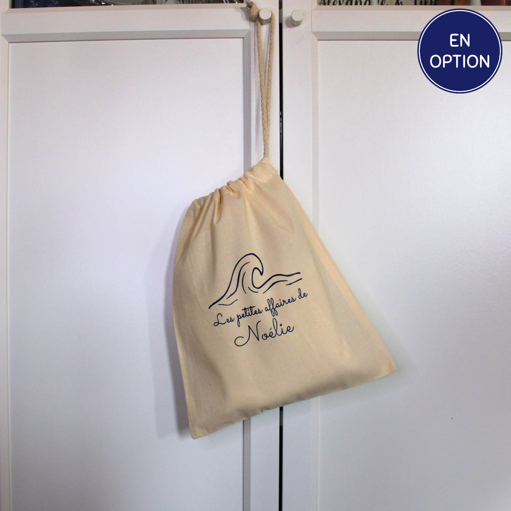 Sac pochon en tissu beige suspendu à une poignée, personnalisé avec l'inscription Les petites affaires de Noélie et un motif de vague.