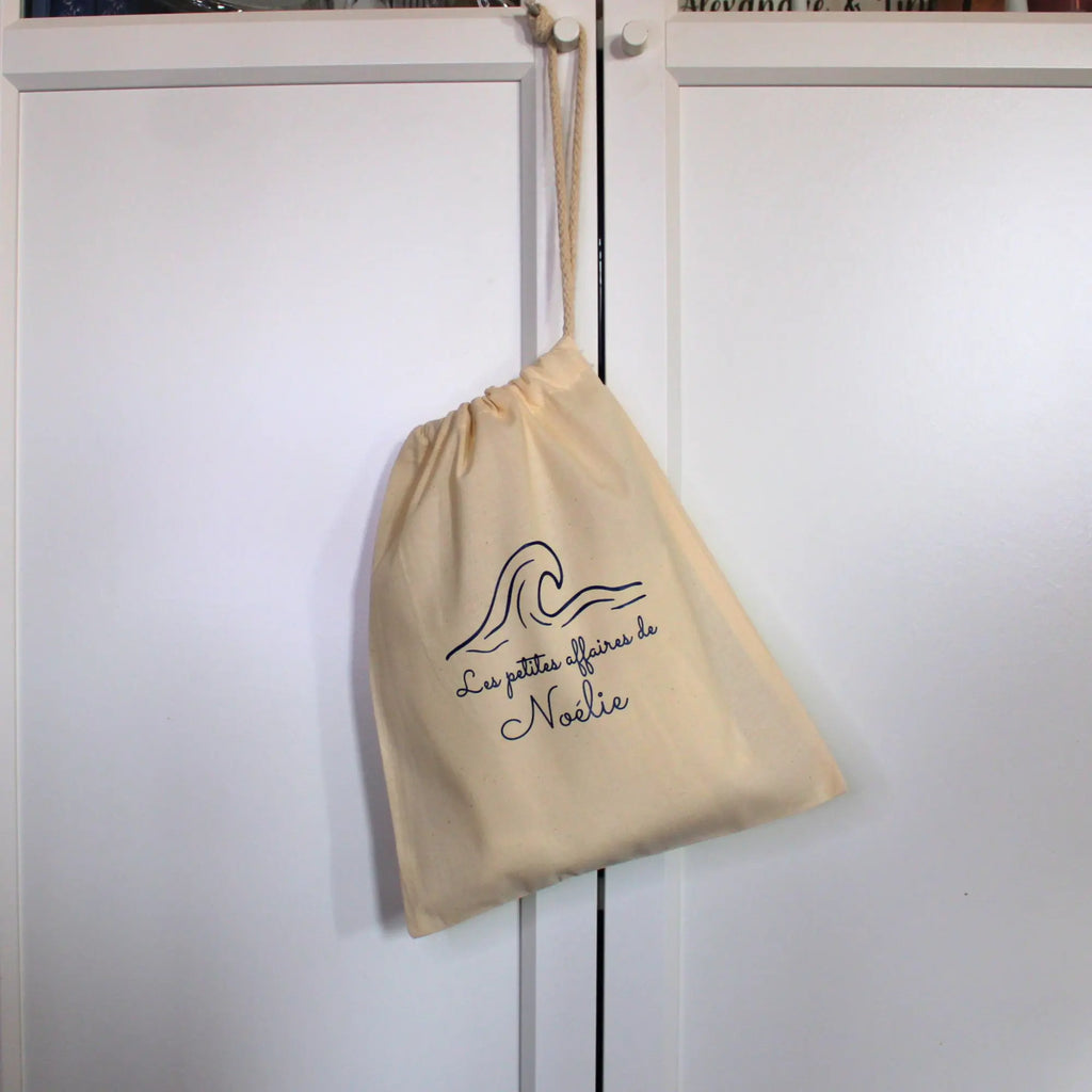 sac pochon en tissu beige suspendu à une poignée, personnalisé avec l'inscription Les petites affaires de Noélie et un motif de vague.