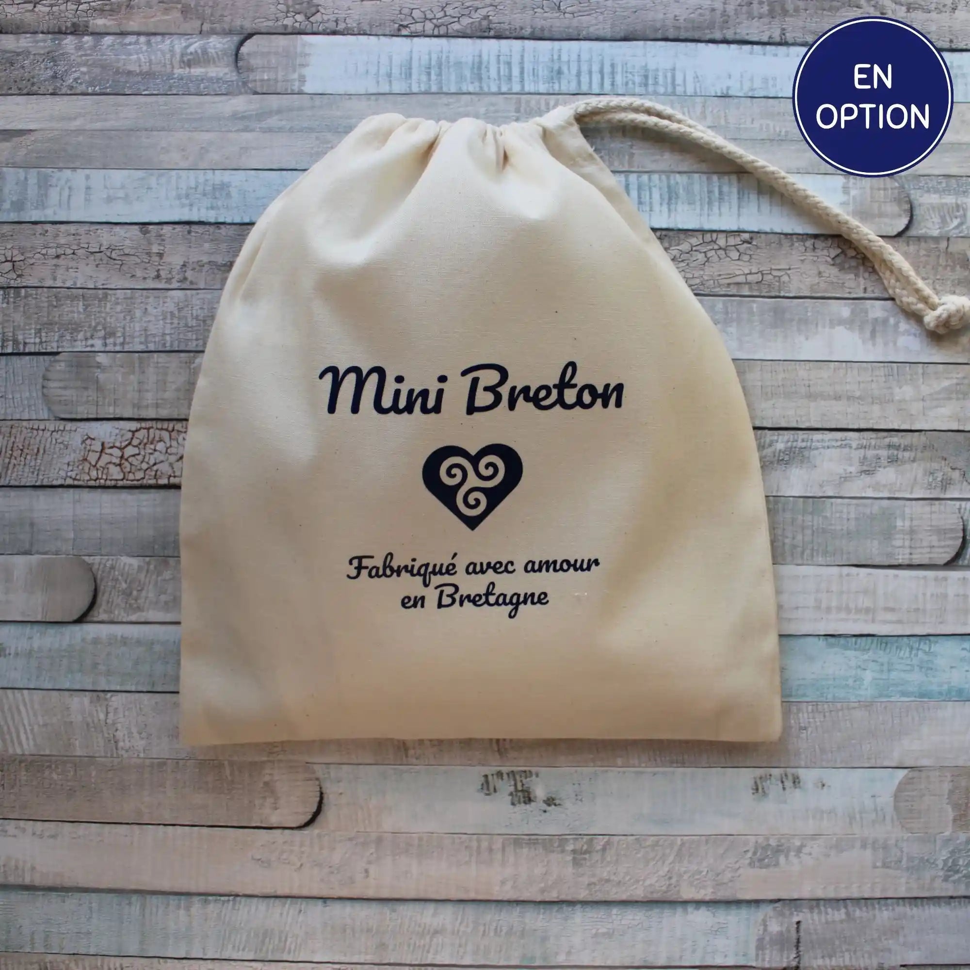 Un petit pochon en toile de coton naturel beige avec un cordon de serrage, orné d'un marquage bleu. L'inscription "Mini Breton" est placée au-dessus d'un symbole cœur celtique breton, suivi de la phrase "Fabriqué avec amour en Bretagne". Le pochon est posé sur un fond en bois clair texturé.