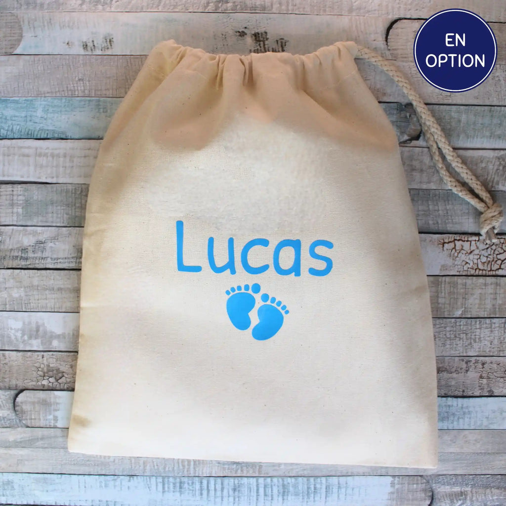 Image en gros plan d'un pochon en coton écru avec cordon, personnalisé avec le prénom "Lucas" en bleu clair et de petites empreintes de pieds